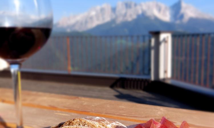 Glas Rotwein neben Brot und Speck auf einem Holztisch, im Hintergrund die Dolomiten