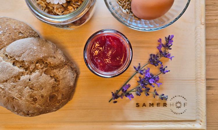 Holzbrett mit Roggenbrot, einem Glas roter Marmelade und Lavendelblüten, im Hintergrund ein Glas Granola und eine Schale mit einem Ei, eingraviertes Samerhof-Logo