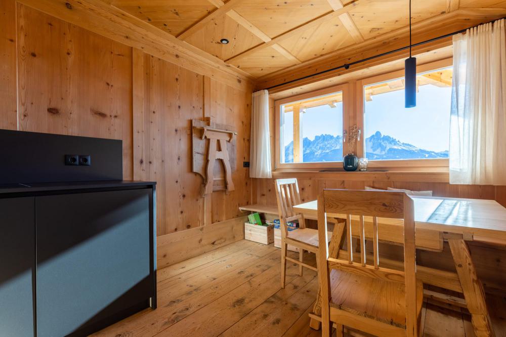 Holzvertäfelter Wohnraum mit Esstisch, Holzstühlen und einer Sitzbank am Fenster, im Hintergrund Blick auf die Dolomiten, links eine dunkle Kochnische Holzvertäfelter Wohnraum mit Esstisch, Holzstühlen und einer Sitzbank am Fenster, im Hintergrund Blick auf die Dolomiten, links eine dunkle Kochnische
