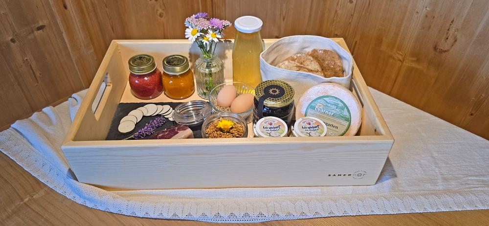 Holzkiste mit Frühstückskiste "Bauernhofgenuss": Marmelade, Honig, Speck, Käse, Eier, Granola, Joghurt, Saft, Brötchen und ein Blumenstrauß, auf einer weißen Tischdecke Holzkiste mit Frühstückskiste "Bauernhofgenuss": Marmelade, Honig, Speck, Käse, Eier, Granola, Joghurt, Saft, Brötchen und ein Blumenstrauß, auf einer weißen Tischdecke