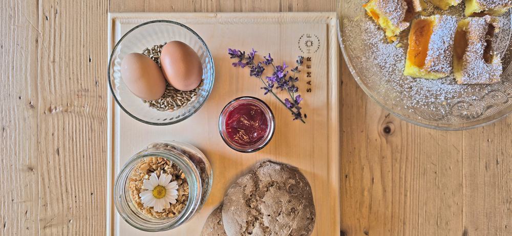 Draufsicht auf ein Holzbrett mit dem Samerhof-Logo: zwei braune Eier in einer Schale mit Sonnenblumenkernen, ein Glas rote Marmelade, ein Glas Granola mit Gänseblümchen, ein Roggenbrötchen und Lavendelblüten, daneben ein Teller mit Marillenkuchen Draufsicht auf ein Holzbrett mit dem Samerhof-Logo: zwei braune Eier in einer Schale mit Sonnenblumenkernen, ein Glas rote Marmelade, ein Glas Granola mit Gänseblümchen, ein Roggenbrötchen und Lavendelblüten, daneben ein Teller mit Marillenkuchen