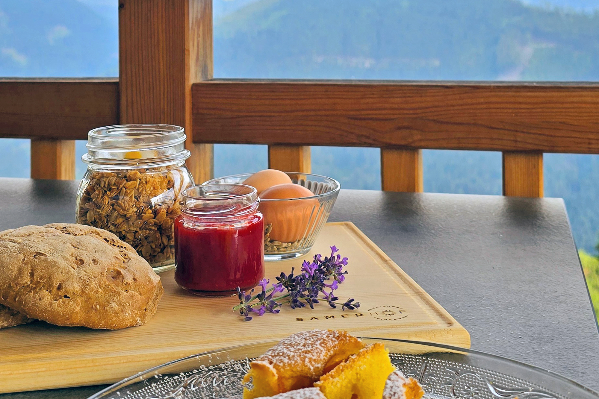 Holzbrett mit Roggenbrötchen, einem Glas Granola, einem Glas roter Marmelade und einer Schale mit zwei braunen Eiern, dekoriert mit Lavendelblüten, im Hintergrund Bergpanorama